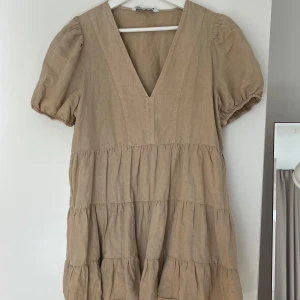 Beige klänning med v-ringning - Säljer en söt beige klänning med v-ringning och korta puffärmar. Klänningen har en volangdetalj och är gjord i ett lätt material, perfekt för varmare dagar. Den har en avslappnad passform och är knälång.