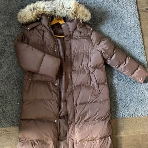 Brun dunjacka med äkta päls - Säljer en helt ny oanvänd lång brun dunjacka med äkta avtagbar päls på huvan. Jackan har långa ärmar och stängs med dragkedja och knappar. Lite tunnare så passar perfekt nu till våren. 