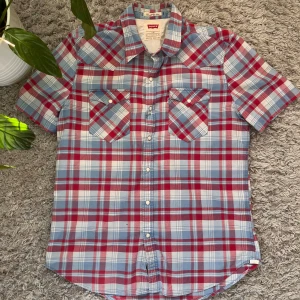 Rutig kortärmad skjorta från Levi's - Säljer en snygg rutig skjorta från Levi's i rött, blått och vitt. Skjortan har korta ärmar och två bröstfickor med knappar. Perfekt för en avslappnad stil. Slim fit för en modern look.