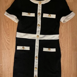 Svart och vit klänning med guldknappar - Elegant svart klänning med vita detaljer och guldknappar. Kortärmad design med dekorativa fickor framtill. Perfekt för en stilren look. Den ger lite gossip girl/old money vibes.