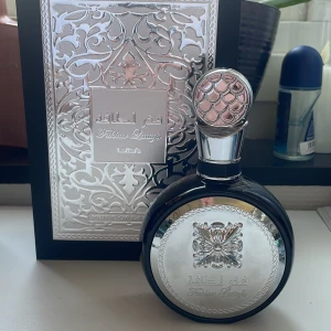 Fakhar Lattafa edp 100ml - Elegant parfym från Lattafa i en rund flaska med silverdetaljer och ett dekorativt mönster. Flaskan har en unik kork med ett fiskfjällsmönster. Förpackningen är stilfullt designad med ett intrikat mönster i silver och svart. Mer än 85ml kvar!