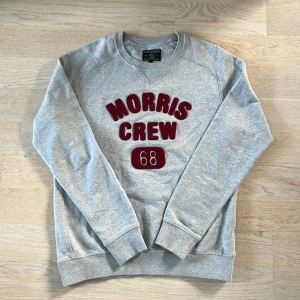 Grå sweatshirt från Morris Gents - Säljer en stilren grå sweatshirt från Morris Gents med röd text 'Morris Crew 68' på framsidan. Tröjan har långa ärmar och en klassisk rund halsringning. Perfekt för en avslappnad stil.