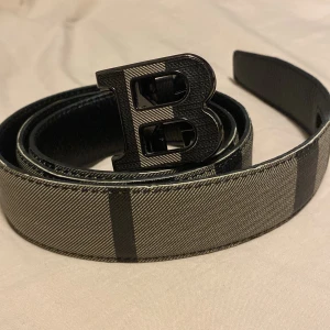 Burberry bälte - Snyggt bälte med ett stort B-spänne i metall. Bältet har ett stilrent randigt mönster i grått och svart, perfekt för att ge en extra touch till din outfit.