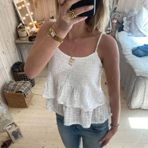 Vit volangtopp - Säljer en söt vit volangtopp med smala axelband och dubbla volanger. Toppen är perfekt för sommardagar och har en luftig passform. Den är gjord i ett lätt och bekvämt material som ger en avslappnad look.
