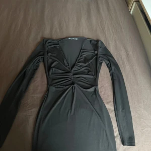 Svart klänning från Nelly  - Snygg svart klänning från Nelly i storlek XS. Klänningen har en v-ringad design med rynkade detaljer framtill och långa ärmar. Perfekt för en stilren look.