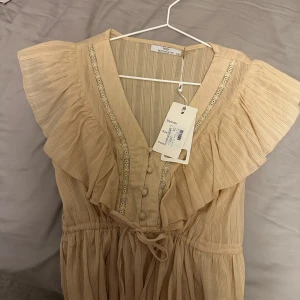 Blus - Säljer en söt beige klänning från Raglady. Blusen har volanger och spetsdetaljer vid axlarna samt knappar framtill. Den är v-ringad och har ett knytband i midjan för en fin passform. Den är helt ny och helt oanvänd. Nytt pris 399kr - mitt pris 299kr