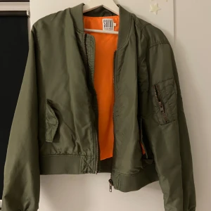 Olivgrön bomberjacka från Saint Tropez - Snygg olivgrön bomberjacka från Saint Tropez med orange foder. Jackan har dragkedja framtill och en ficka med dragkedja på ärmen. Perfekt för en cool och avslappnad stil.