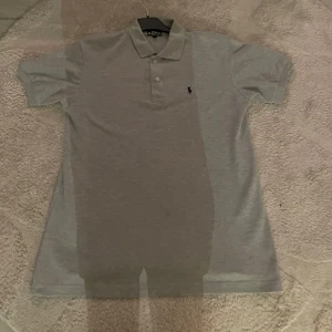 Grå pikétröja från Polo Ralph Lauren - Säljer en stilren grå pikétröja från Polo Ralph Lauren. Tröjan har korta ärmar och en klassisk krage med två knappar. Den ikoniska loggan är broderad på bröstet. Perfekt för en avslappnad men ändå elegant look.