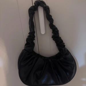 Svart axelväska med rynkad rem - Snygg svart axelväska med en rynkad rem. Väskan har en mjuk och stilren design, perfekt för att ge din outfit en trendig touch. Tillverkad i ett material som liknar skinn.❤️‍🔥