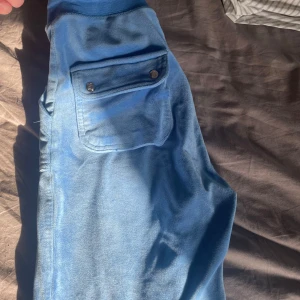 Blå Juicybyxor - Snygga blå byxor med en praktisk ficka med knappar på sidan. Perfekta för en avslappnad stil. Byxorna har en mjuk och bekväm känsla. Storlek XS