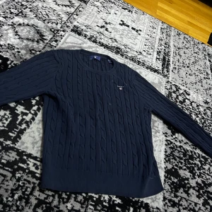 Mörkblå stickad tröja från GANT - Säljer en snygg mörkblå stickad tröja från GANT. Tröjan har ett klassiskt flätmönster och rund halsringning. Perfekt för kyligare dagar. Passar ungdomar i åldern 13-14 år.