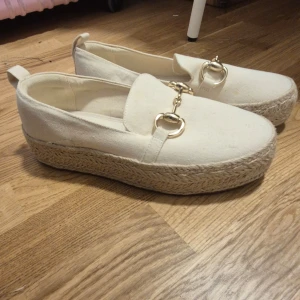 Beige loafers med gulddetaljer - Snygga beige loafers med en hög sula i flätad stil och guldfärgade detaljer på ovansidan. Perfekta för en stilren look med en touch av elegans. De har en bekväm passform och är lätta att ta på.