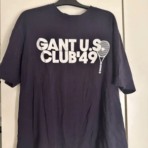 Svart t-shirt från Gant - Säljer en svart t-shirt från Gant med trycket 'GANT U.S. CLUB '49' och en tennisracket på framsidan. T-shirten har korta ärmar och en klassisk rund halsringning. Perfekt för en avslappnad stil. Tröjan är använd fåtal gånger och är sann i storlek💞