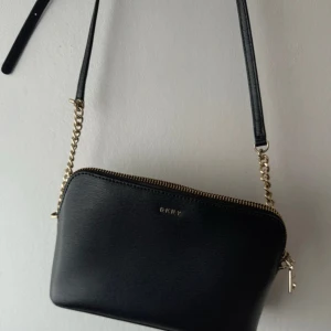  DKNY BRYANT DOME CROSSBODY - Snygg svart axelväska från DKNY med guldfärgad kedjedetalj och dragkedja upptill. Väskan är tillverkad i skinn. Oanvänd. 