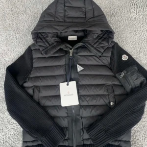 Svart pufferjacka från Moncler - Snygg svart pufferjacka från Moncler med huva och dragkedja. Jackan har stickade ärmar och en quiltad kropp, vilket ger en cool kontrast. Perfekt för kyligare dagar.