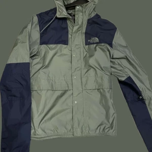 Grå och marinblå vindjacka från The North Face - Säljer en stilren grå och marinblå vindjacka från The North Face. Jackan har en praktisk huva och stängs med knappar. Perfekt för blåsiga dagar och har en klassisk passform.