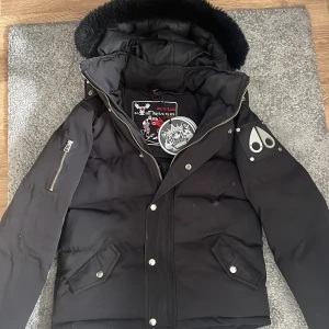 Svart dunjacka från Moose Knuckles - Säljer en svart dunjacka från Moose Knuckles med pälskantad huva och dragkedja. Jackan har långa ärmar och flera fickor med knappar. Perfekt för kalla dagar med stil. 🖤