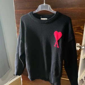 Svart tröja med rött hjärta från Ami Paris - Snygg svart tröja från Ami Paris med ett rött hjärta och bokstaven 'A' på bröstet. Tröjan har långa ärmar och ribbade kanter vid ärmslut och nederkant. Perfekt för en stilren look.