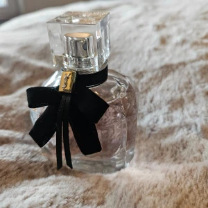 Yves Saint Laurent Mon Paris EdP 50 ml - Elegant parfymflaska från Yves Saint Laurent med en lyxig svart rosett och en fyrkantig kork. Flaskan är genomskinlig och innehåller en ljusrosa doftvätska. Perfekt för den som älskar en sofistikerad och modern stil. Använd max 10 sprut. Inköpt på Kicks.