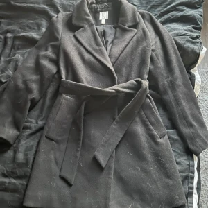 Svart kappa från H&M - Snygg svart kappa från H&M med knytskärp i midjan. Perfekt för kyligare dagar med sin klassiska design och långa ärmar. Ett stilrent val för garderoben.