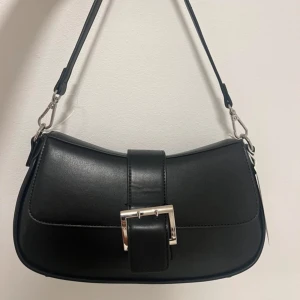 Svart axelväska med spänne - Snygg svart axelväska i skinn med ett stilrent spänne framtill. Väskan har en justerbar axelrem och en enkel design som passar till många tillfällen. Perfekt för att bära dina nödvändigheter med stil.