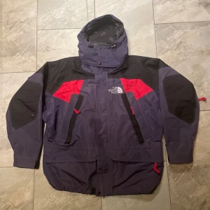 Svart och röd jacka från The North Face - Säljer en cool svart och röd jacka från The North Face. Jackan har en justerbar huva, dragkedja och knappar framtill. Den är långärmad med praktiska fickor och detaljer i rött. Perfekt för kyligare väder! Funkar även jättebra för skid säsongen.