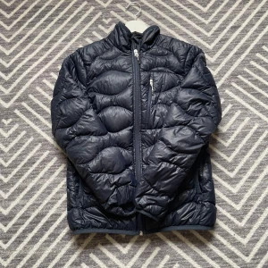 Pufferjacka från Peak Performance - Säljer en snygg mörkblå pufferjacka från Peak Performance. Jackan har en quiltad design med dragkedja framtill och en hög krage. Perfekt för kommande vår dagar och ger en stilren look. Passar både till vardags och mer avslappnade tillfällen.