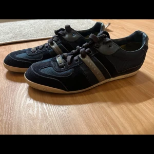 Mörk blåa sneakers från Hugo Boss - Snygga mörkblåa  sneakers från Hugo Boss med grå detaljer och snörning. Skorna har en stilren design med en kombination av mocka och syntetmaterial. Perfekta för en casual look.