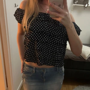 Svart offshoulder topp med prickar - Snygg svart offshoulder topp med vita prickar och volangdetaljer. Perfekt för en somrig look. Toppen är croppad och ger en trendig stil.