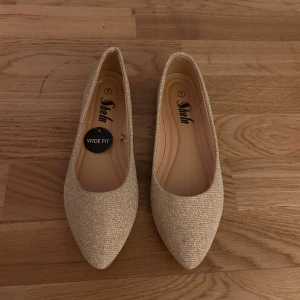 Glittriga ballerinaskor i guld - Snygga glittriga ballerinaskor i guld med spetsig tå och bred passform. Perfekta för att lägga till lite glamour till din outfit. Skorna har en låg klack och är bekväma att bära hela dagen. osäker på storleken i eu storlekar.