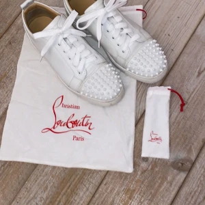 Vita sneakers med nitar från Christian Louboutin - Snygga vita sneakers från Christian Louboutin med ikoniska röda sulor och coola nitar på tån. Skorna har snörning och är tillverkade i skinn, vilket ger en lyxig känsla. Perfekta för att ge din outfit en edgy touch. Mycket bra använda och endast fåtal gånger. Pris går att diskuteras vid snabb affär.