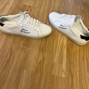 Vita sneakers från Saint Laurent - Snygga vita sneakers från Saint Laurent i skinn. Klassisk design med snörning och en diskret logga. Tillverkade i Italien, dessa skor kombinerar stil och komfort perfekt. Skicket är 7/10 då det är lite creases men inte så farligt. Självklart tvättar jag de innan man får med allt i bilden 2 påsar och extra snöre och skorna. Äkta! De är i 40,5 men passar 41 