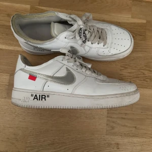 Nike Air Force 1 x Off-White - Säljer ett par använda vita Nike Air Force 1 x Off-White sneakers med den ikoniska silver swooshen och textdetaljer. Skorna har snörning med texten 'SHOELACES' och den klassiska 'AIR' texten på sulan. Perfekta för den som vill ha en unik touch på sina sneakers.  (Obs repclicor) 