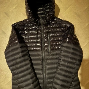 Svart dunjacka från Moose Knuckles - Säljer en svart pufferjacka från Moose Knuckles. Jackan har en glansig finish med dragkedja framtill och en huva. Den är lätt och tillverkad av återvunnet material, perfekt för kyliga dagar. Märkeslogga på ärmen ger en extra touch.