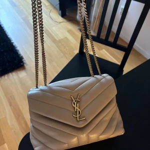 Saint Laurent väska  - Snygg beige axelväska från YSL med quiltad design och kedjeaxelrem. Väskan har en elegant YSL-logga i guld på framsidan och är perfekt för att ge din outfit en lyxig touch. Den kommer med kvitto och order bekräftelse. 