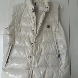Vit dunväst från Moncler - Säljer en snygg vit dunväst från Moncler med hög krage och dragkedja. Västen har knappar och två praktiska fickor med dragkedja. Perfekt för kyliga dagar när du vill hålla stilen. Strl 3 (M)