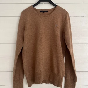 Brun stickad tröja från GANT - Säljer en stilren brun tröja från GANT med rund hals och ribbade muddar. Perfekt för en avslappnad och snygg look. Tröjan har en liten broderad logga på bröstet. Mvh GrischBolaget