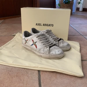Vita sneakers från Axel Arigato - Säljer ett par vita sneakers från Axel Arigato med broderade fåglar i flera färger på sidan. Skorna har snörning och en klassisk design som passar perfekt för en stilren look.