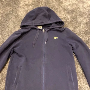 Mörkblå hoodie från Lacoste - Säljer en snygg mörkblå hoodie från Lacoste med dragkedja och klassisk krokodillogga på bröstet. Hoodien har långa ärmar och en justerbar huva med snören. Perfekt för en avslappnad stil.