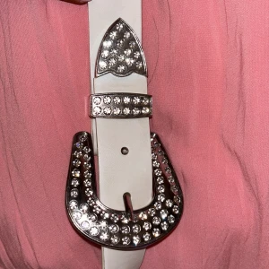 Vitt bälte med glittrigt spänne - Snyggt vitt bälte med ett stort, glittrigt spänne dekorerat med strass. Perfekt för att ge en extra touch till din outfit. Bältet har en klassisk design med en modern twist.