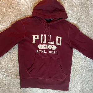 Vinröd hoodie från Ralph Lauren - Säljer en snygg vinröd hoodie från Ralph Lauren med texten 'POLO 1967 ATHL. DEPT.' på framsidan. Tröjan har en klassisk känguruficka och justerbar huva. Perfekt för en avslappnad stil. Nyskick inte mycket använd!