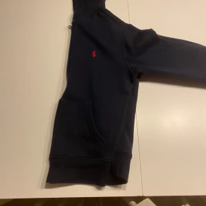 Mörkblå hoodie från Ralph Lauren - Säljer en snygg mörkblå hoodie från Ralph Lauren med dragkedja och det klassiska röda logotypbroderiet på bröstet. Perfekt för en avslappnad stil. Hoodien har långa ärmar och en praktisk ficka framtill.
