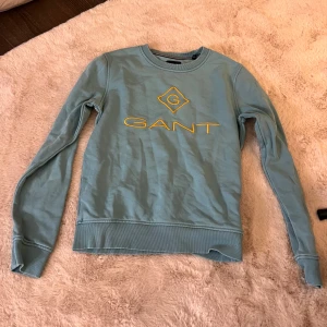 Blå tröja från GANT - Säljer en snygg blå tröja från GANT med gul logga på bröstet. Tröjan har långa ärmar och ribbade muddar vid ärmslut och nederkant. Perfekt för en avslappnad stil.