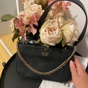 Svart axelväska från Guess - Snygg svart axelväska från Guess med guldfärgad kedjedetalj och dragkedja på baksidan. Perfekt för att ge din outfit en elegant touch. Väskan har en stilren design och är lätt att matcha med olika stilar.