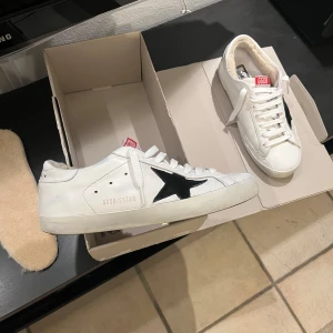 Golden Goose superstar - Snygga vita sneakers från Golden Goose med en svart stjärna på sidan. Skorna har snörning och en röd detalj på plösen. Insidan är fodrad med ett mjukt material för extra komfort. Perfekta för en stilren look. I fint skick. Nya snören men original snören med kommer. Fås även med extra sulor som är fodrade. Skriv järna vid funderingar!