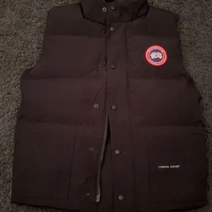 Svart dunväst från Canada Goose - Säljer en svart dunväst från Canada Goose med deras ikoniska märke på bröstet. Västen har en dragkedja och knappar framtill samt praktiska fickor. Perfekt för kyliga dagar. Har liten effek vid armen men inget man märker när man eller nån annan märker när man har på den 