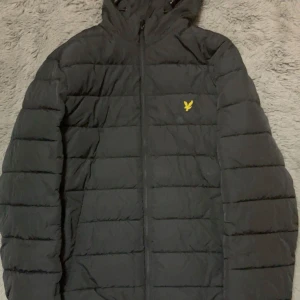 Svart pufferjacka från Lyle & Scott - Säljer en svart pufferjacka från Lyle & Scott med huva och dragkedja. Jackan har ett quiltat mönster och en gul logotyp på bröstet. Perfekt för kyligare dagar.