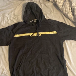 Svart hoodie från Boss - Säljer en stilren svart hoodie från Boss med en guldfärgad rand och text på bröstet. Den har en klassisk huva med dragsko och långa ärmar. Perfekt för en avslappnad stil. Funkar som L eftersom den är ganska tajt 