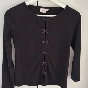 Svart ribbad topp från Nelly - Säljer en stilren svart ribbad topp från Nelly med långa ärmar och söta rosett-detaljer framtill. Perfekt för en chic och enkel look. Passar bra till både jeans och kjol.