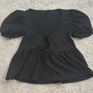 Svart blus med puffärmar - Säljer en svart blus med puffiga kortärmar och knytning framtill. Blusen har en v-ringad design och är tillverkad i ett ribbat material som ger en fin struktur. Perfekt för en stilren look.